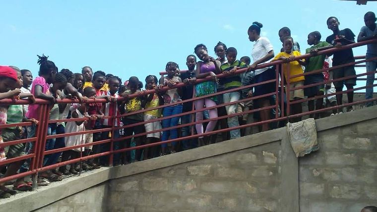 Crowd of people standing behind a red railing on a concrete bridge, looking down under a bright sky
