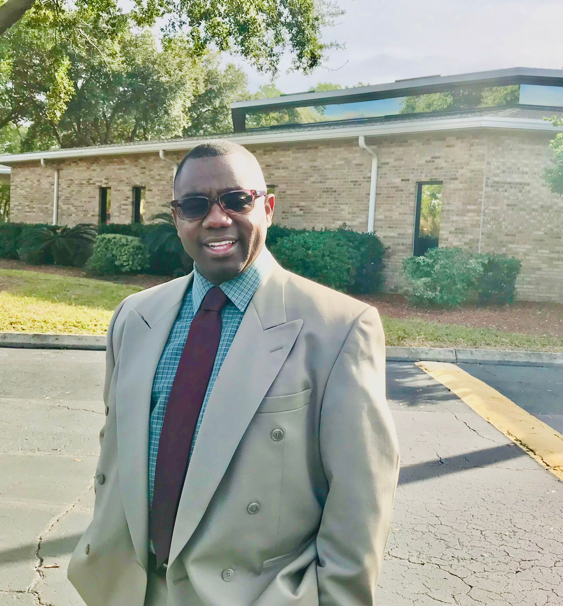 Man in a beige suit and sunglasses standing outdoors in front of a brick building and trees