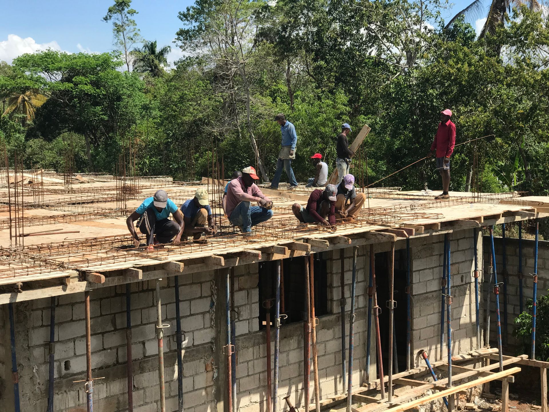 Workers pour concrete on a roof under construction, with trees and blue sky in the background.