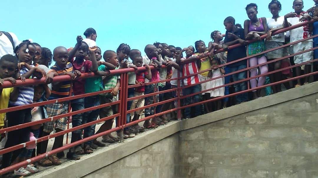 Crowd of people standing behind a red railing on a concrete bridge, looking down outdoors.