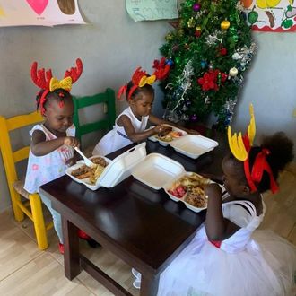 Children in festive headbands eating lunch at a small table beside a decorated Christmas tree